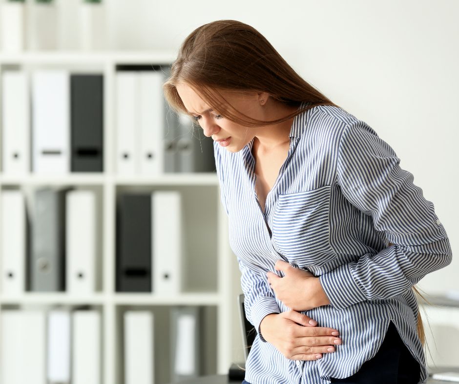 Woman with brown hair wearing a striped shirt, holding her stomach in pain with a bookcase in the background